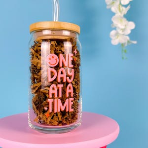 Może przedstawiać: Szklany słoik z bambusową pokrywką i słomką. Słoik ma różowy wzór z napisem "One Day At A Time" i uśmiechniętą buźką.