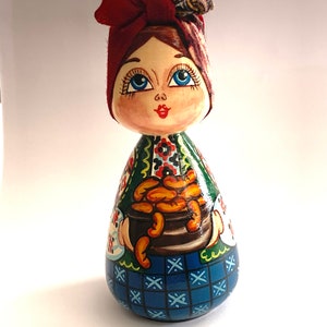 以下が含まれることがあります： 赤いリボン、青い目、緑と青のドレスを着た手描きの木製人形。人形はオレンジ色の果物の入ったバスケットを持っています。