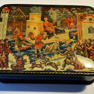 Op de afbeelding: Een houten doos met een zwarte gelakte afwerking en een gouden rand. Het deksel heeft een kleurrijk geschilderd tafereel van mensen en dieren in een winterse setting. De scène bevat een groep mensen in traditionele kleding, een paardenslee en een hond.