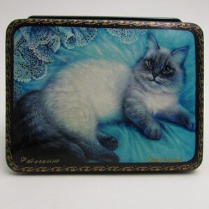 Puede incluir: Una caja rectangular negra con una escena pintada de un gato esponjoso blanco y gris de ojos azules descansando sobre una superficie de tela azul brillante con detalles de encaje blanco. El borde tiene adornos dorados y texto cirílico.