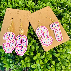 May include: A pair of donut-shaped earrings with sprinkles. The earrings are in the shape of the numbers 39 and 21. The earrings are on a brown card with a hole in the top.