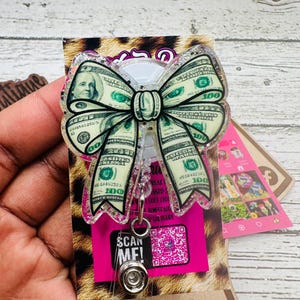May include: A clear badge reel with a bow-shaped design featuring a $100 bill pattern. The bow is outlined with glitter and has a pink backing with the text "SCAN ME!".