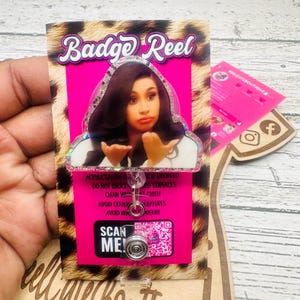 Könnte beinhalten: Ein pinkfarbenes und leopardenmusterndes Badge-Reel mit einem Prominentenbild. Das Reel hat ein einziehbares Kabel und einen Metallclip. Der Text "Badge Reel" steht oben und der Text "Scan Me!" steht auf der Vorderseite.
