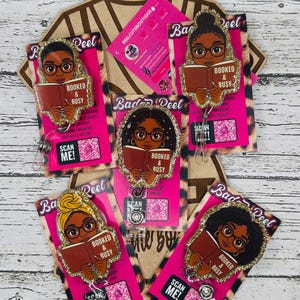 May include: A collection of "Booked & Busy" badge reels featuring illustrations of Black women reading books. Each reel has a glittery border and is attached to a pink card with the text "Scan Me!". The reels are displayed on a leopard print background.