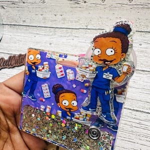 May include: A badge reel with a cartoon character in a blue uniform, holding a bottle of pills. The background is filled with pills and bottles. The badge reel has a glittery border and the words "Pharmacy Tech" are visible.