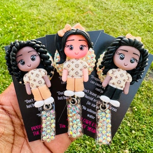May include: Three small dolls with black hair and brown eyes wearing a beige and brown patterned dress with a white bow. The dolls are holding a small clear rectangle with rhinestones. The text on the cards reads "Don't leave your doll in humid or cloudy areas", "Don't hot/humid", "Don't hot/humid", "No submersion in water", "Care extra of your doll", "Dropping can cause your doll to reel/pull apart", "The doll is made of polymer clay and is not waterproof."