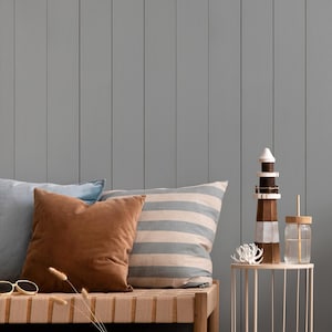 May include: A coastal-themed interior scene with a gray paneled wall. A wooden bench with pillows, a round rattan chair, and a small side table with decorative items, including a wooden lighthouse and a glass with a straw.