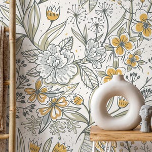 Op de afbeelding: Crèmekleurig bloemenbehang met grote grijze, gele en groene bloemen. Een witte vaas met een cirkelvormige opening, een kleine grijze buste en een decoratief object staan op een houten tafel. Een bamboeladder en een beige deken staan links.