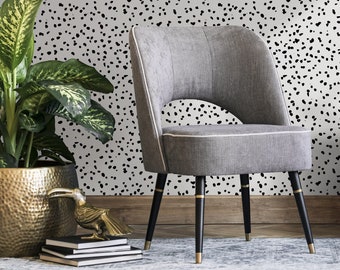 Puede incluir: Una silla tapizada gris con patas negras y detalles dorados se encuentra frente a una pared blanca con un patrón de lunares negros y blancos.