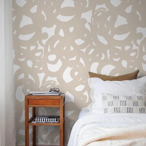 May include: Bedroom scene featuring a wooden nightstand with a drawer and two shelves. The wall is adorned with a beige and white abstract pattern. A bed with white linens and pillows is visible, creating a cozy atmosphere.