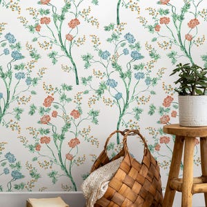 May include: White floral wallpaper with watercolor-style branches bearing orange, blue, and yellow flowers. A woven basket with a blanket sits on a wooden floor. A small potted plant and a wooden stool are also present.