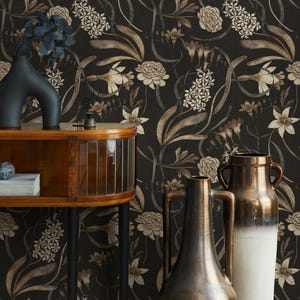 Puede incluir: Papel pintado floral oscuro con diseños botánicos crema y marrón. Un mueble de madera con puerta de cristal curvada y patas negras está en primer plano, junto con varios jarrones decorativos en negro, marrón y blanco.