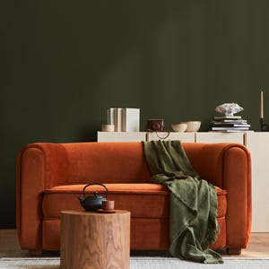 May include: An orange velvet sofa with rounded arms and a wooden side table. A green throw blanket is draped over the sofa. A black teapot and a small cup sit on the table. A white cabinet with decorative items is in the background.
