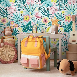 K&ouml;nnte beinhalten: Ein gelber Rucksack mit rosa Taschen h&auml;ngt an einem Holzstuhl vor einer wei&szlig;en Wand mit tropischem Muster. Ein Stofftier-Affe sitzt in einem Weidenkorb und ein braunes Stofftier-Hund liegt auf dem Boden.