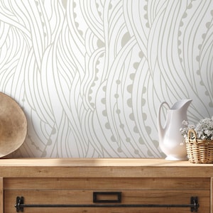 May include: A white ceramic pitcher and a small woven basket with white flowers sit on a wooden surface. A straw hat rests on the left. The background features a white wall with a beige, flowing, abstract pattern.