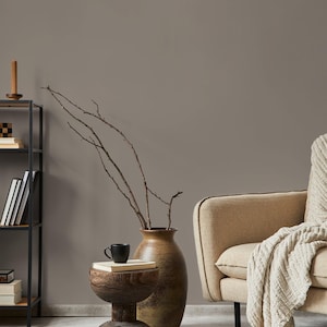 Könnte beinhalten: Gemütliches Wohnzimmer mit neutraler Farbpalette. Ein hölzerner Beistelltisch mit einer schwarzen Tasse und einem Buch, neben einer großen Vase mit Zweigen. Ein beiger Sessel mit einer cremefarbenen Decke befindet sich auf der rechten Seite. Ein schwarzes Metallregal und eine Kerze sind links.