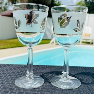 Op de afbeelding: Twee heldere wijnglazen met matglas en zilveren bladeren. De glazen hebben een gouden band en staan op een heldere steel en voet. De achtergrond toont een zwembad en groen.