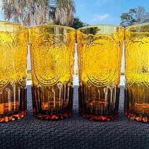 May include: Four amber-colored drinking glasses with ornate embossed designs. The glasses have a tall, cylindrical shape and a rich, warm hue. The embossed pattern is visible on the front of each glass, adding a touch of elegance. The glasses are suitable for serving beverages.