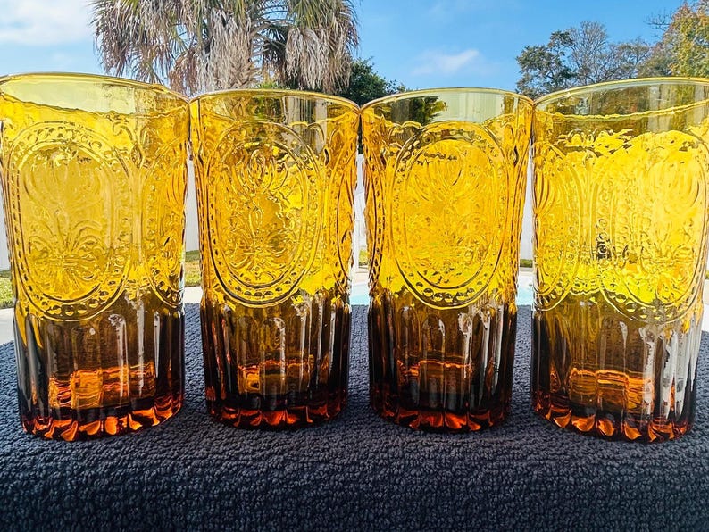May include: Four amber-colored drinking glasses with ornate embossed designs. The glasses have a tall, cylindrical shape and a rich, warm hue. The embossed pattern is visible on the front of each glass, adding a touch of elegance. The glasses are suitable for serving beverages.