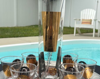 May include: A set of seven clear glass shot glasses with gold coin designs and a clear glass pitcher with gold coin designs. The pitcher has a silver metal stirrer inside. The glasses and pitcher are on a silver tray with a dark wood pattern.
