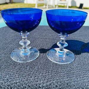 May include: Two cobalt blue champagne coupe glasses. The bowls are a deep blue, transitioning to clear stems and bases. The glasses are displayed on a textured, dark gray surface, with a pool and outdoor seating in the background.