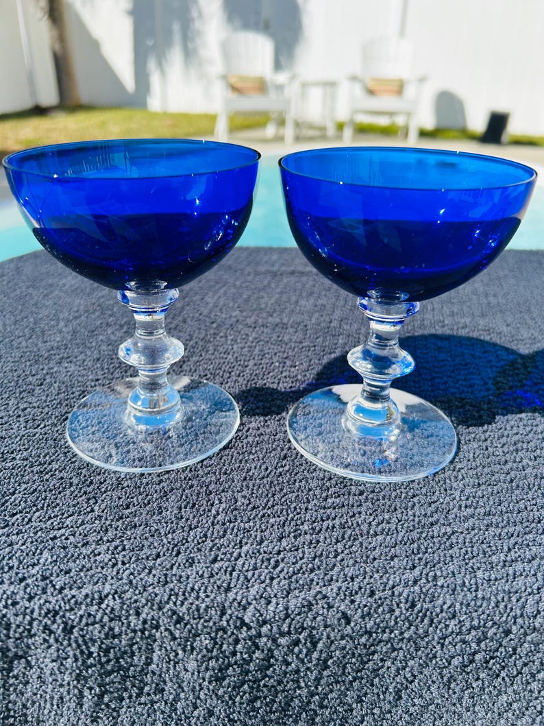 May include: Two cobalt blue champagne coupe glasses. The bowls are a deep blue, transitioning to clear stems and bases. The glasses are displayed on a textured, dark gray surface, with a pool and outdoor seating in the background.