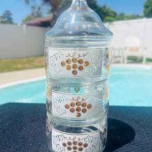 Puede incluir: Un tarro de vidrio transparente de tres niveles con un remate dorado. Cada nivel presenta una etiqueta blanca con un diseño de uvas doradas. El tarro está diseñado para almacenamiento y exhibición.
