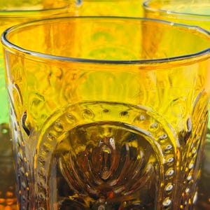 May include: Close-up of several amber-colored drinking glasses. The glasses have a detailed, embossed design around the middle and a smooth rim. The glasses are arranged closely together, with the focus on the front glass.