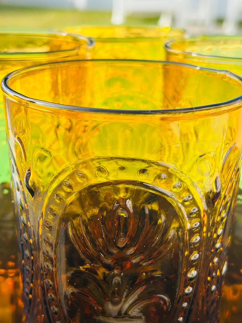 May include: Close-up of several amber-colored drinking glasses. The glasses have a detailed, embossed design around the middle and a smooth rim. The glasses are arranged closely together, with the focus on the front glass.