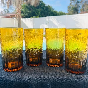 May include: Four amber-colored drinking glasses with embossed floral designs. The glasses are tall and cylindrical, with a slightly flared base. They are arranged in a row on a dark blue surface, with a pool and trees in the background.