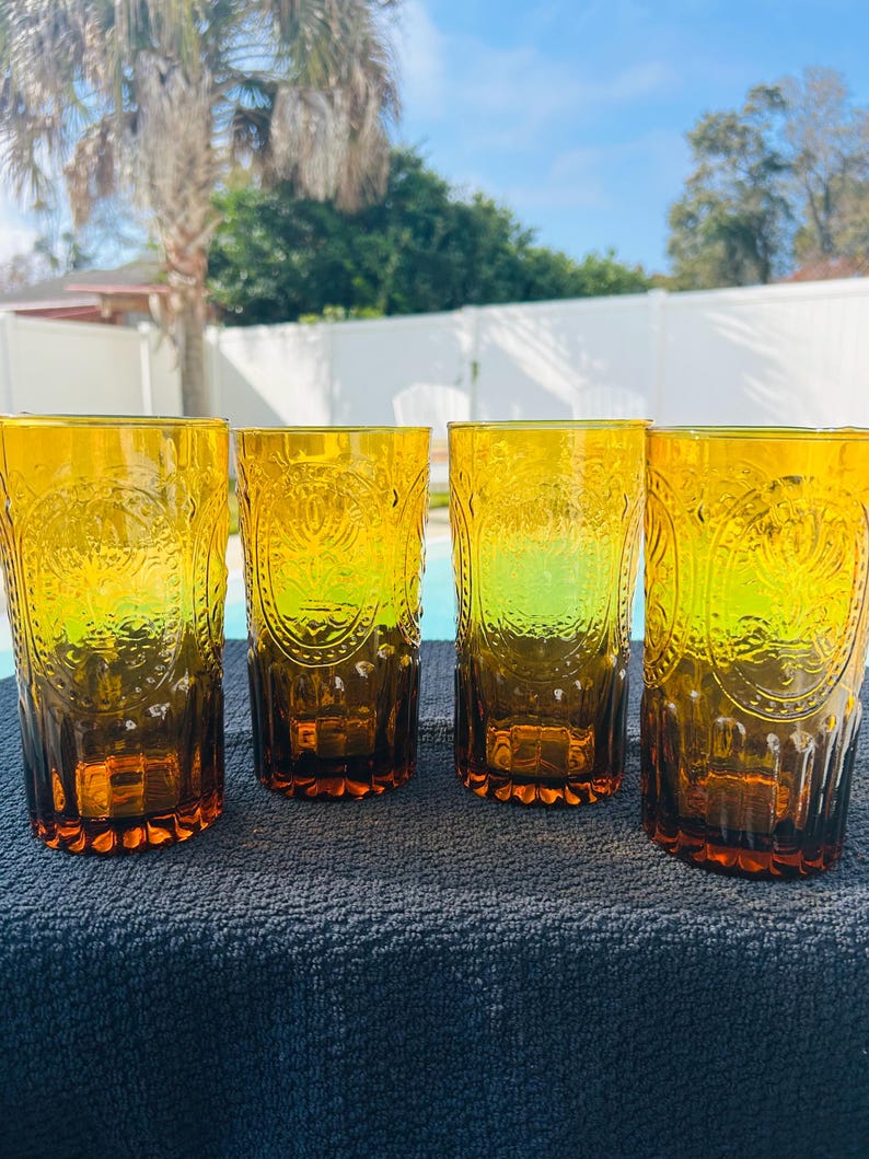 May include: Four amber-colored drinking glasses with embossed floral designs. The glasses are tall and cylindrical, with a slightly flared base. They are arranged in a row on a dark blue surface, with a pool and trees in the background.