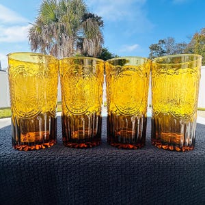 May include: Four amber-colored drinking glasses with a decorative embossed design. The glasses have a tall, cylindrical shape with a textured pattern and a darker amber base. The glasses are arranged in a row, showcasing their vintage style.