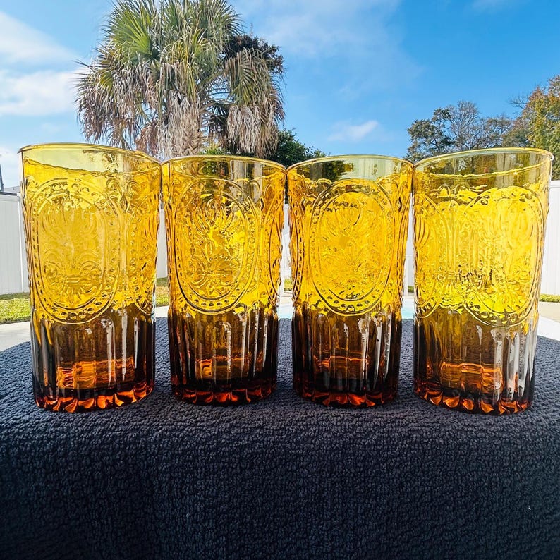 May include: Four amber-colored drinking glasses with a decorative embossed design. The glasses have a tall, cylindrical shape with a textured pattern and a darker amber base. The glasses are arranged in a row, showcasing their vintage style.