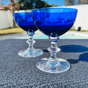 May include: Two cobalt blue stemmed glasses with clear stems and bases. The glasses have a rounded bowl shape. The glasses are sitting on a dark blue textured surface, with a bright blue sky in the background.