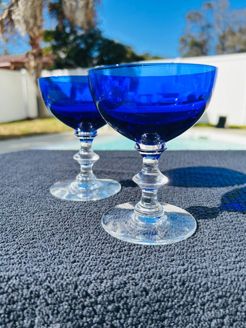 May include: Two cobalt blue stemmed glasses with clear stems and bases. The glasses have a rounded bowl shape. The glasses are sitting on a dark blue textured surface, with a bright blue sky in the background.