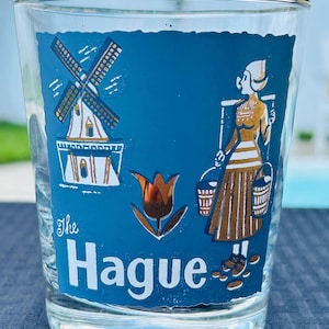 May include: A blue glass with a gold rim and a gold design of a windmill, a tulip, and a woman in traditional Dutch clothing. The text "The Hague" is printed on the glass.