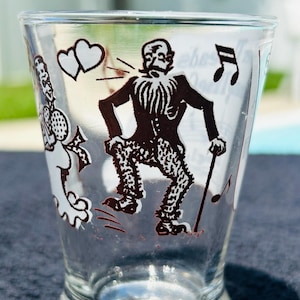 May include: A clear glass tumbler featuring a black graphic print of a bearded figure in a suit and patterned trousers holding a cane. The design includes two hearts, musical notes, and a partial figure on the left side with a microphone.