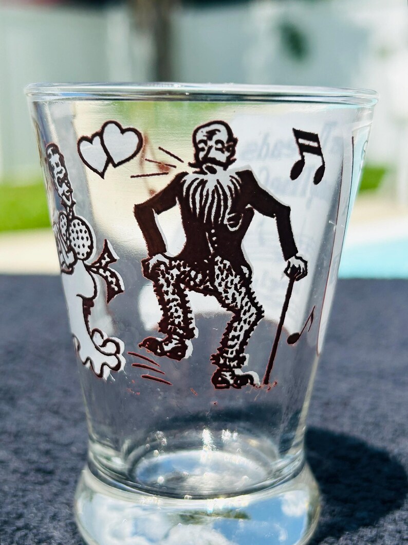 May include: A clear glass tumbler featuring a black graphic print of a bearded figure in a suit and patterned trousers holding a cane. The design includes two hearts, musical notes, and a partial figure on the left side with a microphone.