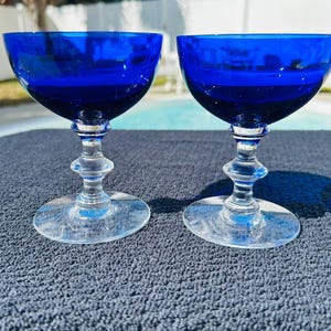 May include: Two cobalt blue coupe glasses with clear stems and bases. The glasses have a rounded bowl shape and are set on a dark blue textured surface. The background includes a swimming pool and trees.