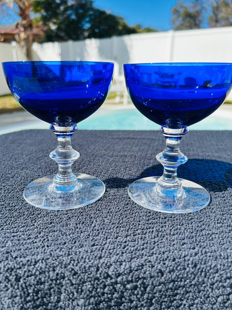 May include: Two cobalt blue coupe glasses with clear stems and bases. The glasses have a rounded bowl shape and are set on a dark blue textured surface. The background includes a swimming pool and trees.