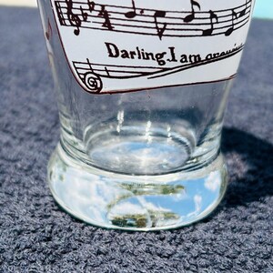 May include: A clear glass tumbler featuring black printed musical staff lines, musical notes, and the text Among The Gold and Darling I am growing. The glass has a thick, rounded base and a tapered shape, with the design printed on the exterior surface.