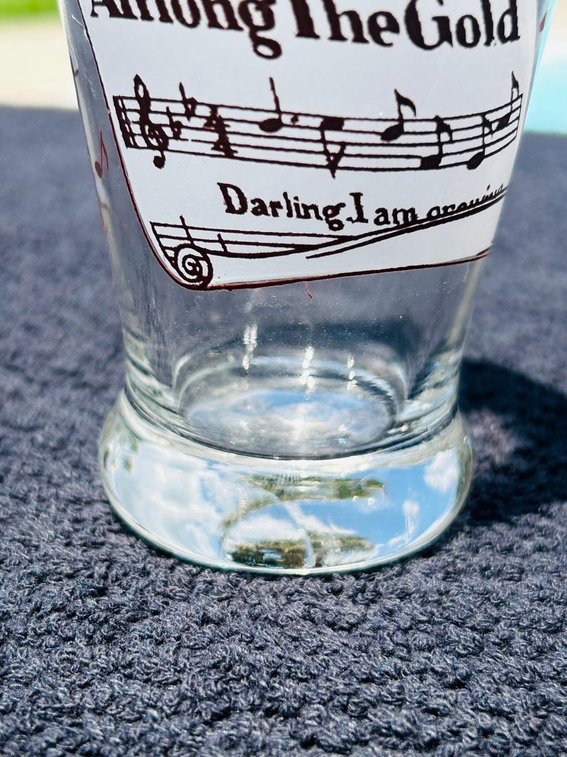 May include: A clear glass tumbler featuring black printed musical staff lines, musical notes, and the text Among The Gold and Darling I am growing. The glass has a thick, rounded base and a tapered shape, with the design printed on the exterior surface.