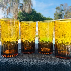 May include: Four amber-colored drinking glasses with an embossed design. The glasses have a cylindrical shape with a dark brown base and a decorative pattern around the middle. The glasses are arranged in a row on a dark surface.