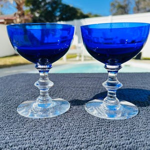 May include: Two cobalt blue champagne coupe glasses. The bowls are a deep blue, contrasting with the clear, ornate stems and bases. The glasses are set against a textured, dark blue surface, with a blurred outdoor background.