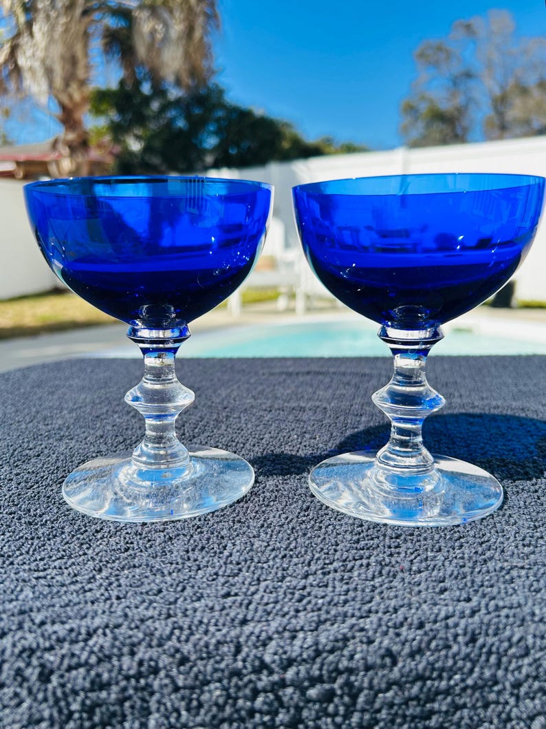 May include: Two cobalt blue champagne coupe glasses. The bowls are a deep blue, contrasting with the clear, ornate stems and bases. The glasses are set against a textured, dark blue surface, with a blurred outdoor background.