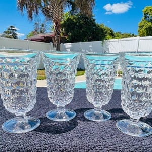 May include: Four clear, stemmed drinking glasses with a diamond pattern. The glasses are displayed on a dark blue surface, with a swimming pool and greenery in the background. The glasses are approximately 7 inches tall.