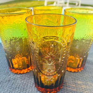 May include: Set of four amber-colored drinking glasses. Each glass features an embossed design with a floral motif and a textured base. The glasses have a vintage aesthetic and are suitable for serving beverages.