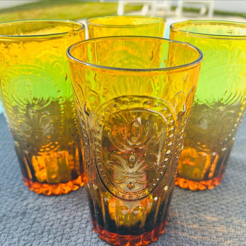 May include: Set of four amber-colored drinking glasses. Each glass features an embossed design with a floral motif and a textured base. The glasses have a vintage aesthetic and are suitable for serving beverages.