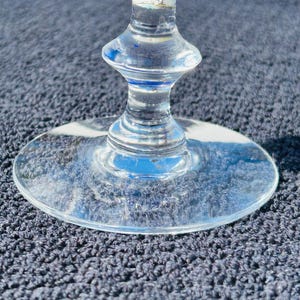 May include: A clear glass wine glass with a blue and white design on the stem. The base of the glass is round and flat. The glass is sitting on a dark blue textured surface.