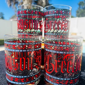 Könnte beinhalten: Satz von vier Vintage-Trinkgläsern. Jedes Glas hat ein rot-schwarzes Buntglasdesign mit dem Wort "Bitter" in gotischer Schrift. Die Gläser haben einen klaren Boden und ein blaues Akzentband.